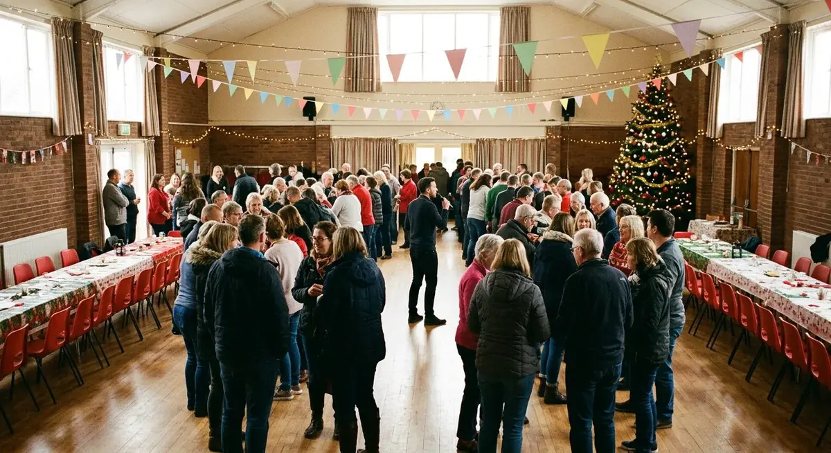 A crowded community hall decorated with bunting, fairy lights and a large Christmas tree, with rows of tables and red chairs pushed to the sides and groups of people gathered in the open space between for a large-group party game.