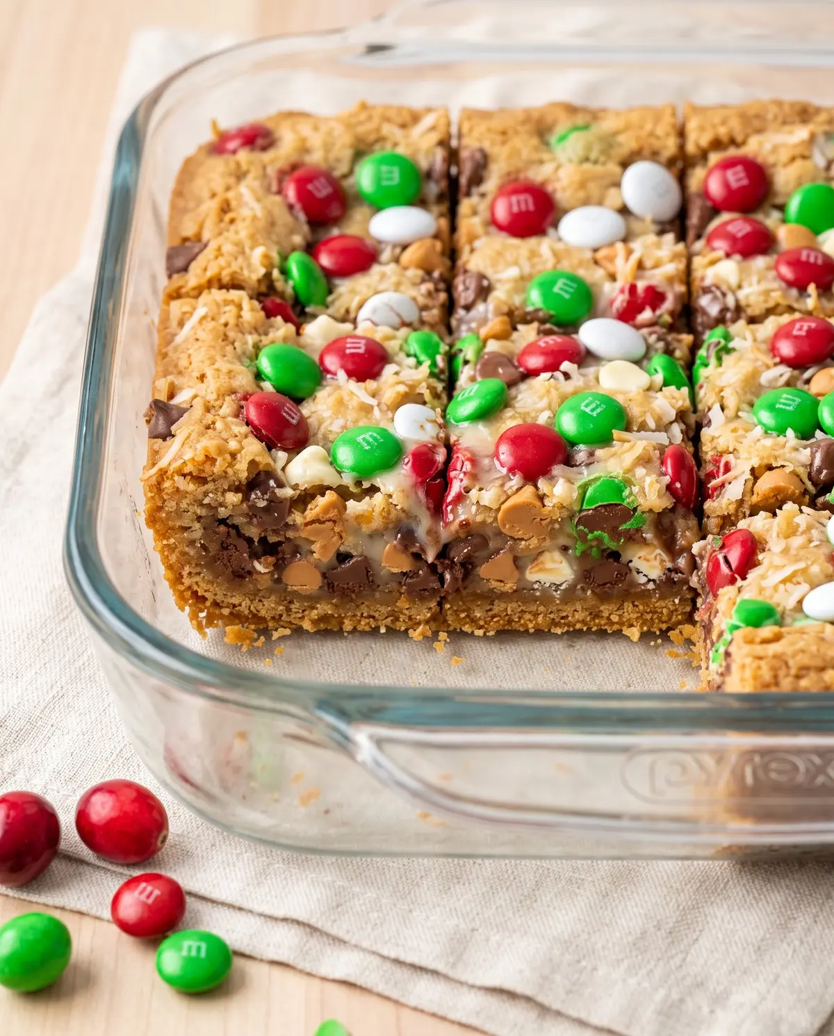 Christmas magic cookie bars in a glass baking dish, viewed from the side, garnished with fresh cranberries