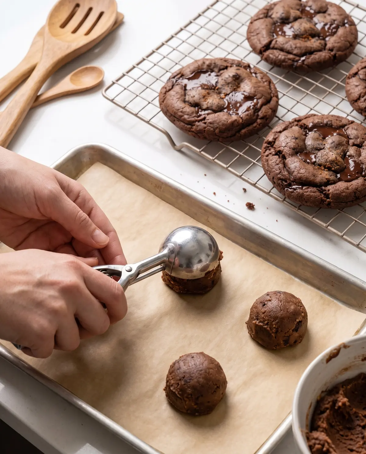 Hands scooping cookie dough balls onto parchment-lined baking sheet with cooling rack nearby
