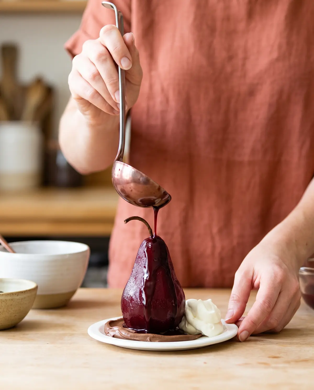 A woman ladles glossy red-wine chocolate sauce over a poached pear