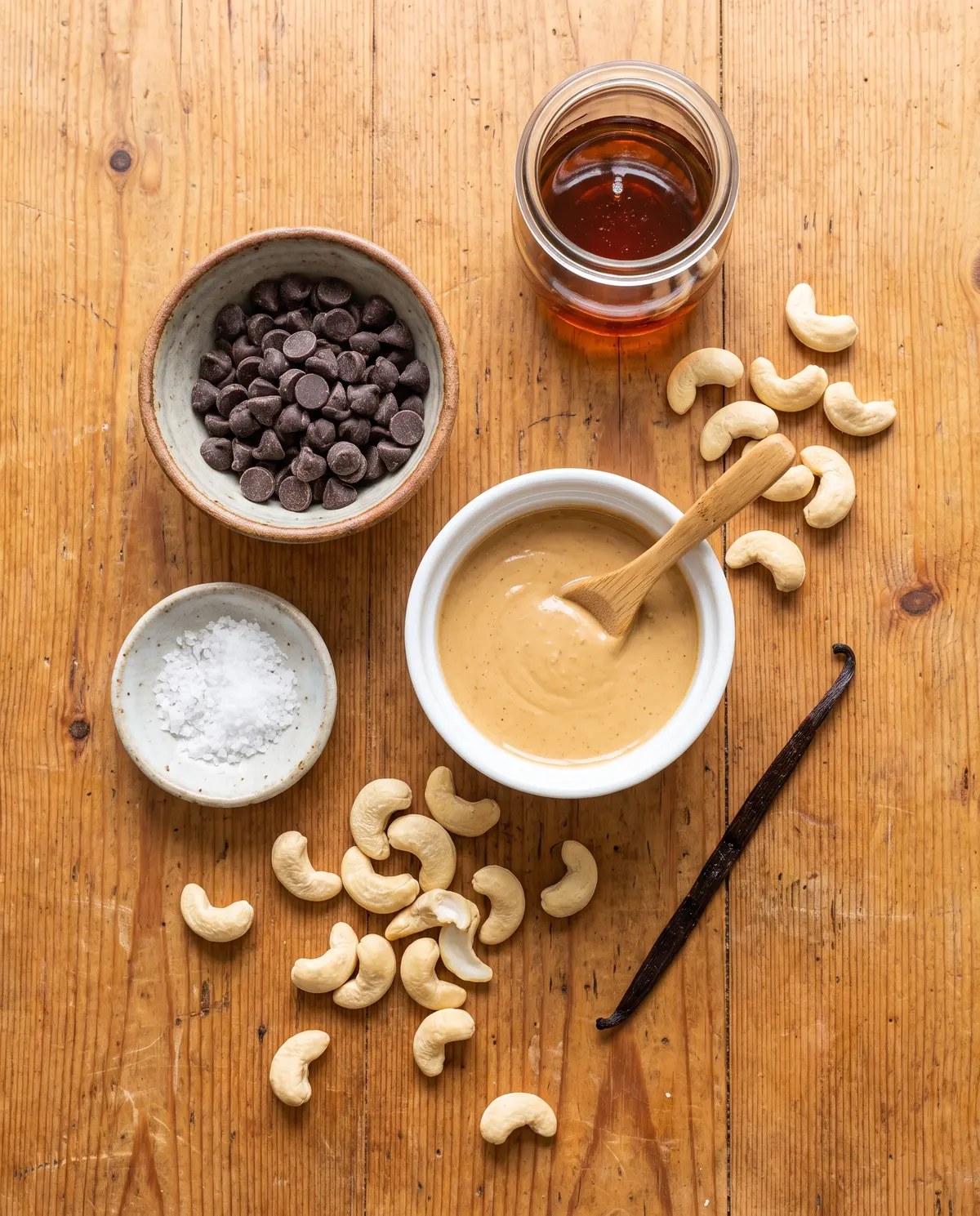 Top-down flat-lay of cashew butter cup ingredients on warm wood: dark chocolate chips, cashew butter, maple syrup, vanilla bean, sea salt, cashews