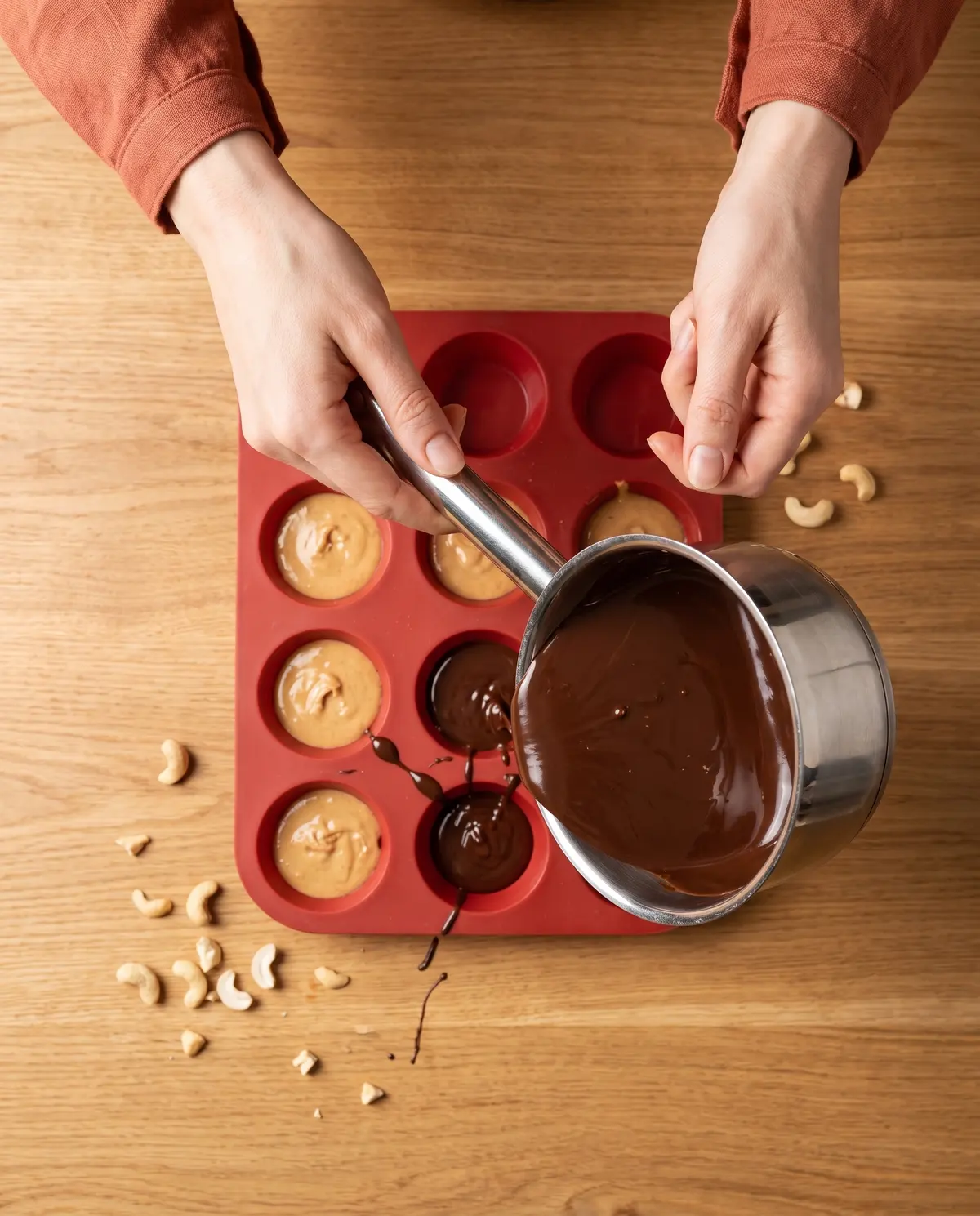 Process shot of feminine hands pouring melted dark chocolate from a small saucepan into a red silicone mold, cashew butter filling visible in some cups, terracotta cuff at the wrist