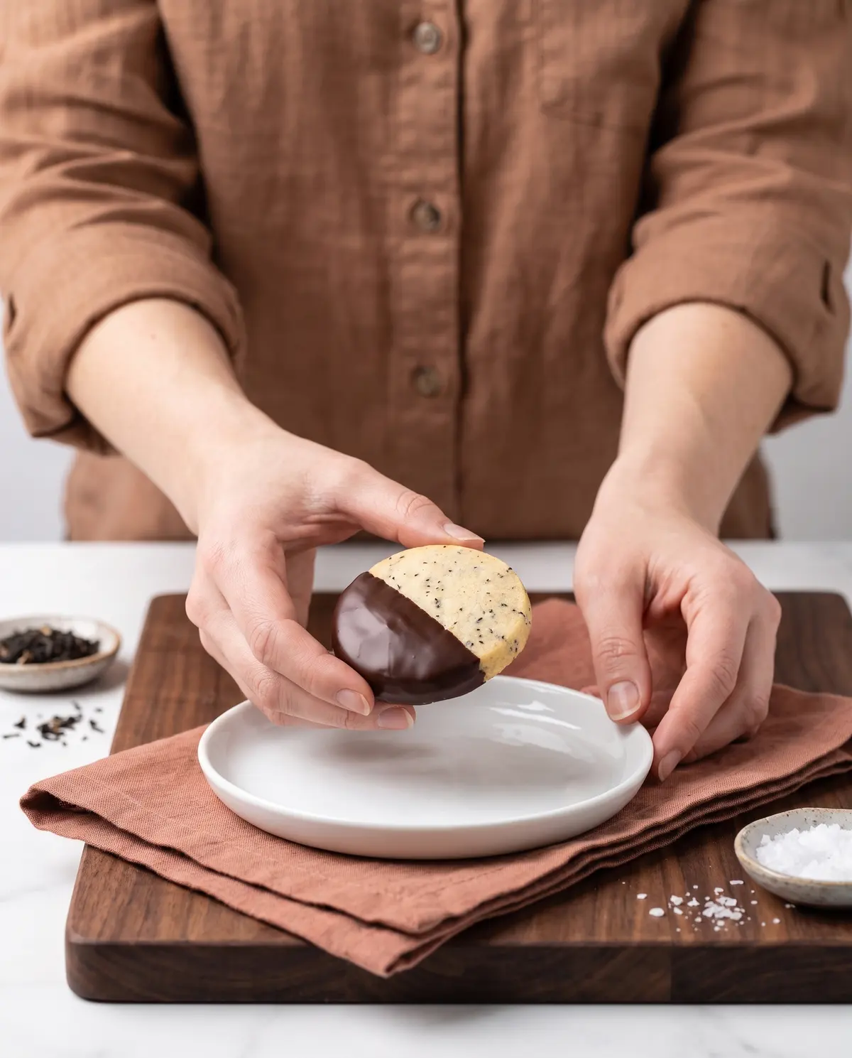A woman's hand holding a freshly dipped chocolate-dipped Earl Grey shortbread cookie above a white plate
