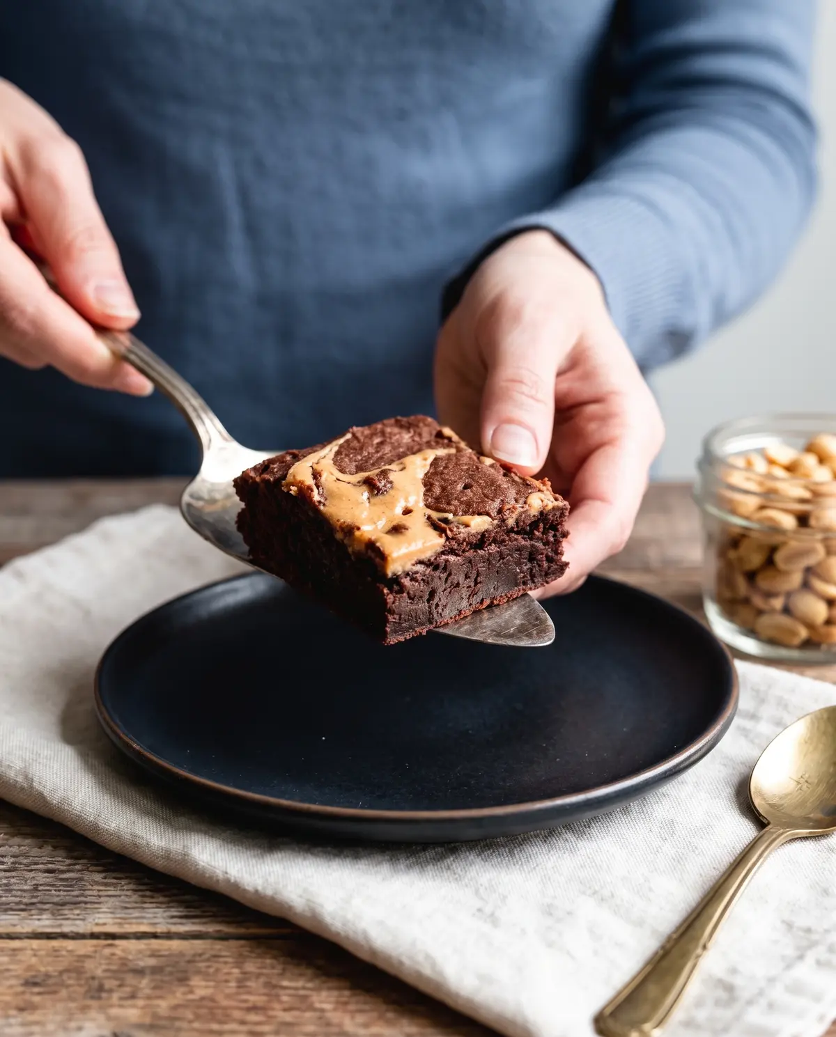 A single fudgy chocolate peanut butter swirl brownie being lifted on a vintage cake server from a dark ceramic plate, dusty blue sleeve visible