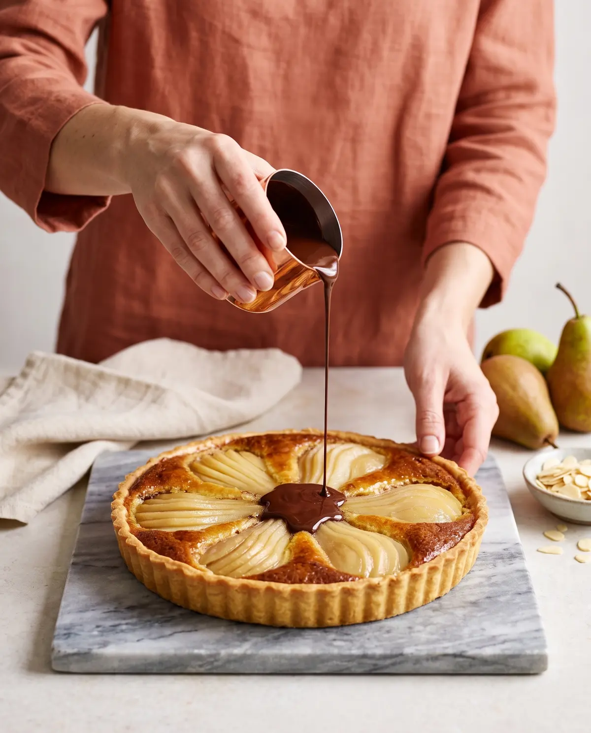A woman's hands pouring melted dark chocolate in a thin stream over the baked tart, wearing a terracotta linen top