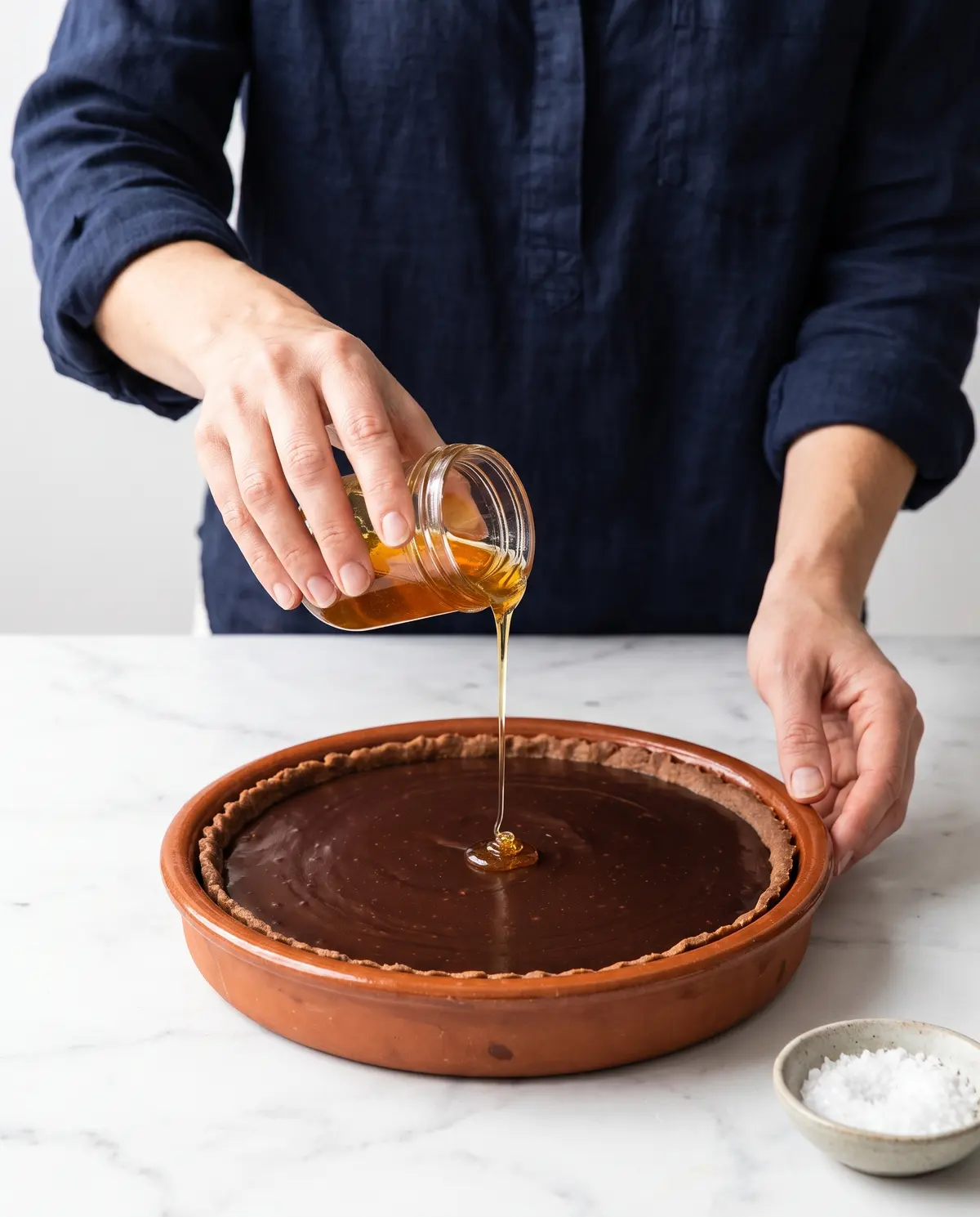 A woman's hands pouring golden honey over a chocolate ganache tart in a terracotta dish