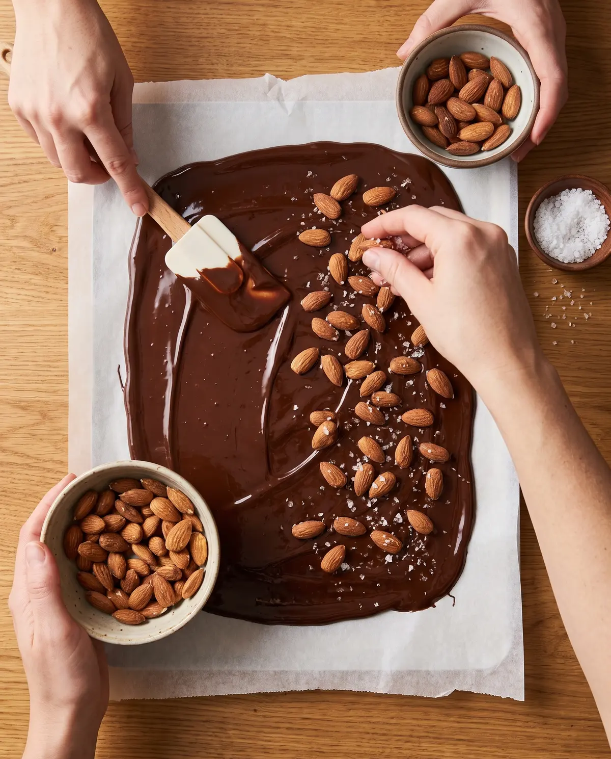 A hand spreading melted chocolate and sprinkling almonds on parchment paper