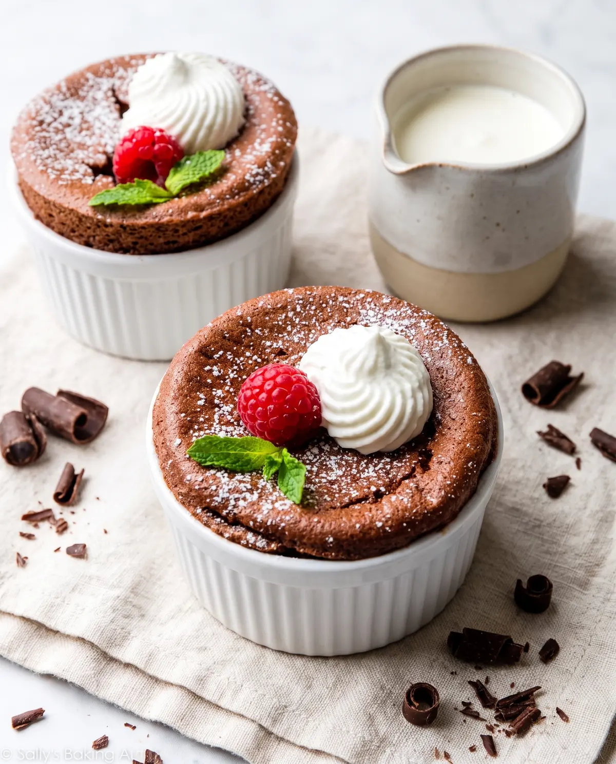 3/4 overhead view of chocolate souffles in white ramekins on a linen cloth with cream and chocolate curls