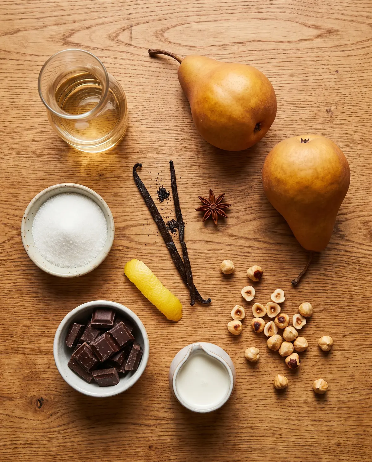 Top-down flat-lay of poached pear ingredients on warm wood: Bosc pears, white wine carafe, sugar, vanilla bean, star anise, lemon zest, dark chocolate, cream, hazelnuts