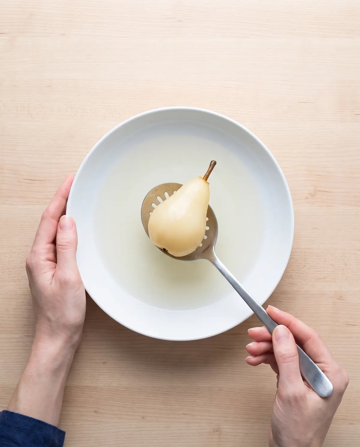 Overhead process shot of feminine hands lifting a poached pear out of a white bowl of white wine syrup with a slotted spoon, navy cuff visible at left wrist edge