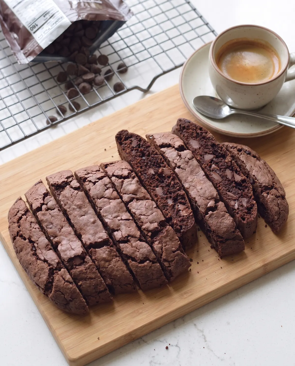 Double chocolate biscotti on cutting board in arc with chocolate chips and cooling rack in background