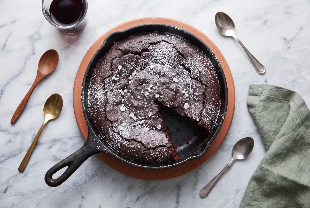 Top-down view of dark chocolate olive oil cake in a cast iron skillet on a terracotta trivet, with a glass of red wine and sage napkin