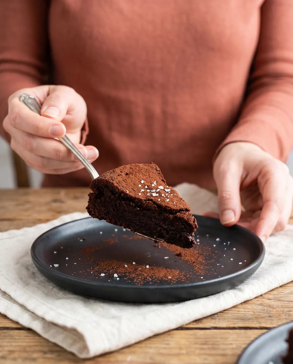 A slice of dark chocolate olive oil cake being lifted on a fork from a dark ceramic plate, terracotta sleeve visible